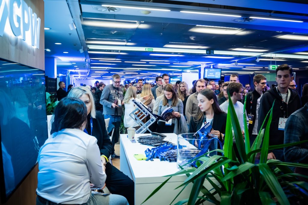 ForFin 2025 - strefa wystawców. Tłum uczestników konferencji finansowej oglądający produkty i usługi finansowe na stanowisku wystawcy przy nowoczesnym białym stole, niebieskie oświetlenie, rozmowy jeden-na-jeden z przedstawicielami instytucji